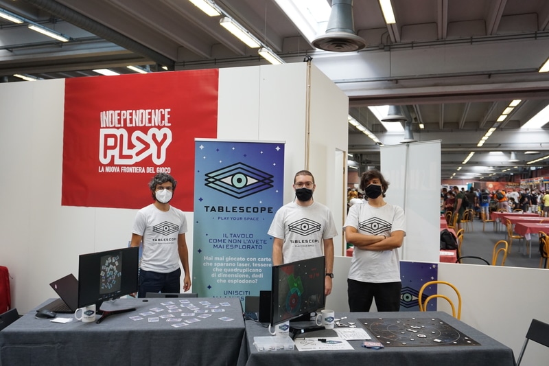 Three boys stand in front of a table in a fair hall. Board game prototypes are on the tables.