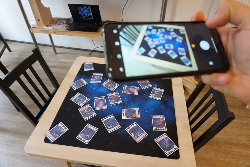 One hand holds up a smartphone, which takes a picture of a table on which cards depicting spaceships are spread out