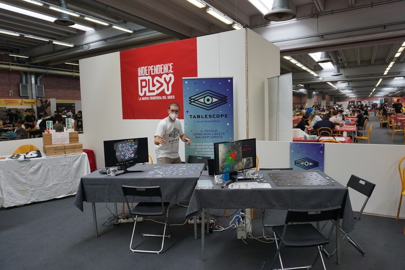 A boy in a dynamic pose behind two tables on which screens and board games are laid