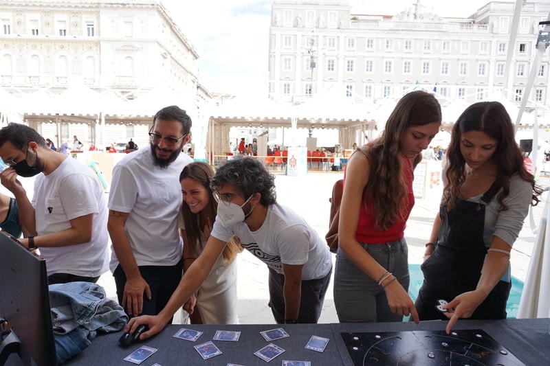 Due tavoli con sopra due schermi e due giochi da tavolo. 5 ragazzi e ragazze giocano divertiti